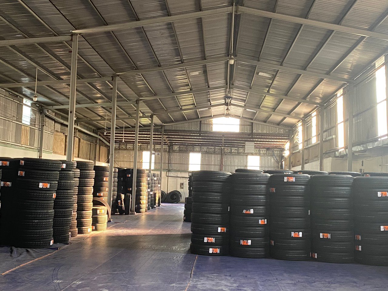 Ảnh chụp kho hàng carmall Tyre chứa nhiều lốp xe xếp thành hàng, trần nhà lợp tôn sáng, sàn nhà màu xanh tím